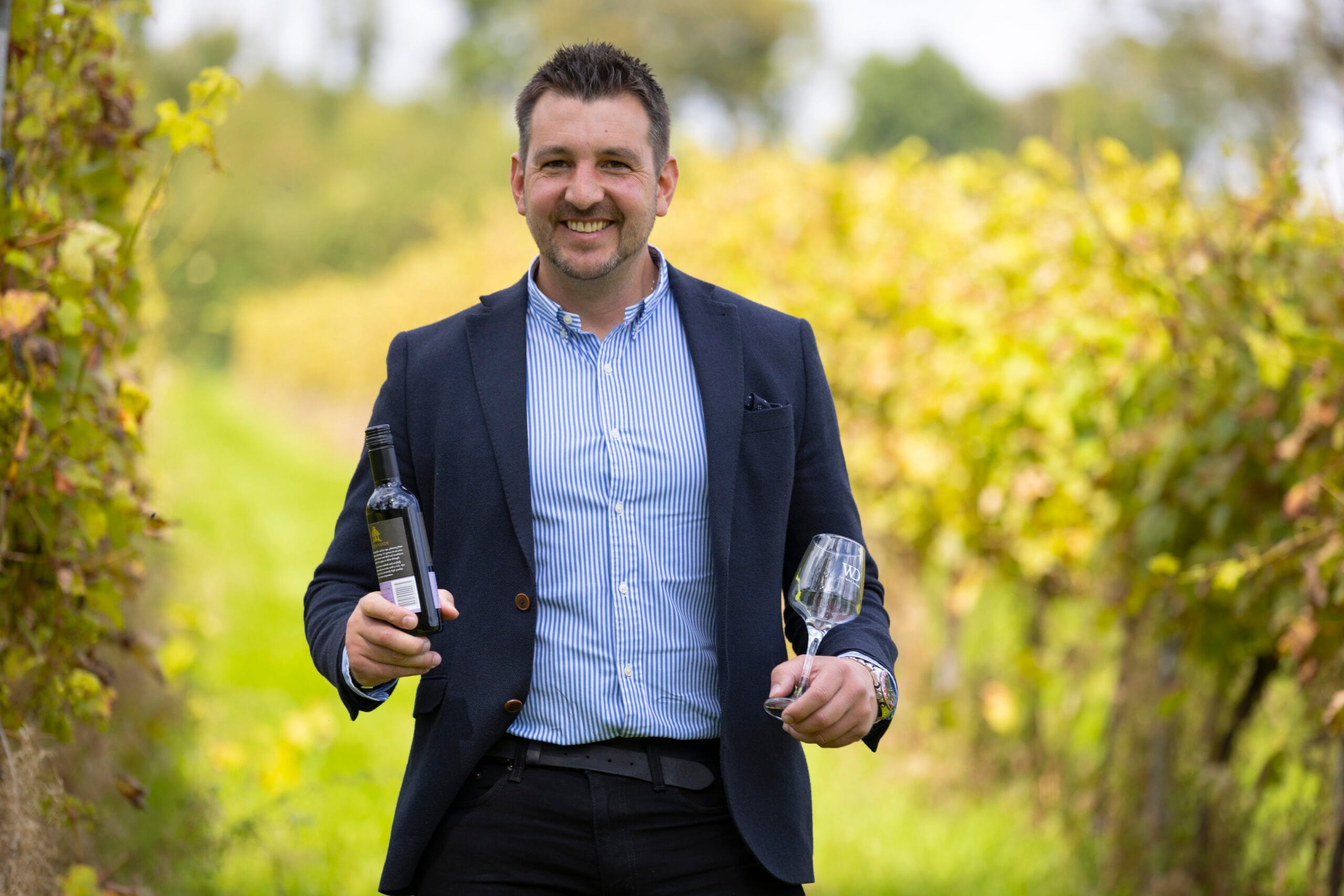 Man in a blazer holding a wine bottle in one hand and a wine glass in the other, standing in a vineyard.