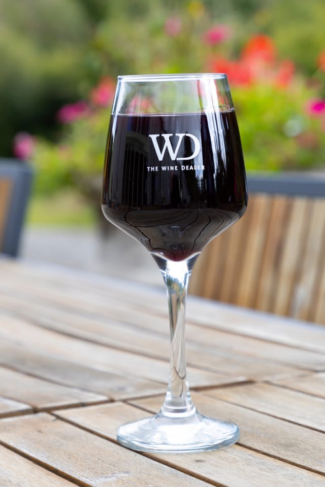 A clear wine glass filled with red wine, featuring the logo of The Wine Dealer.